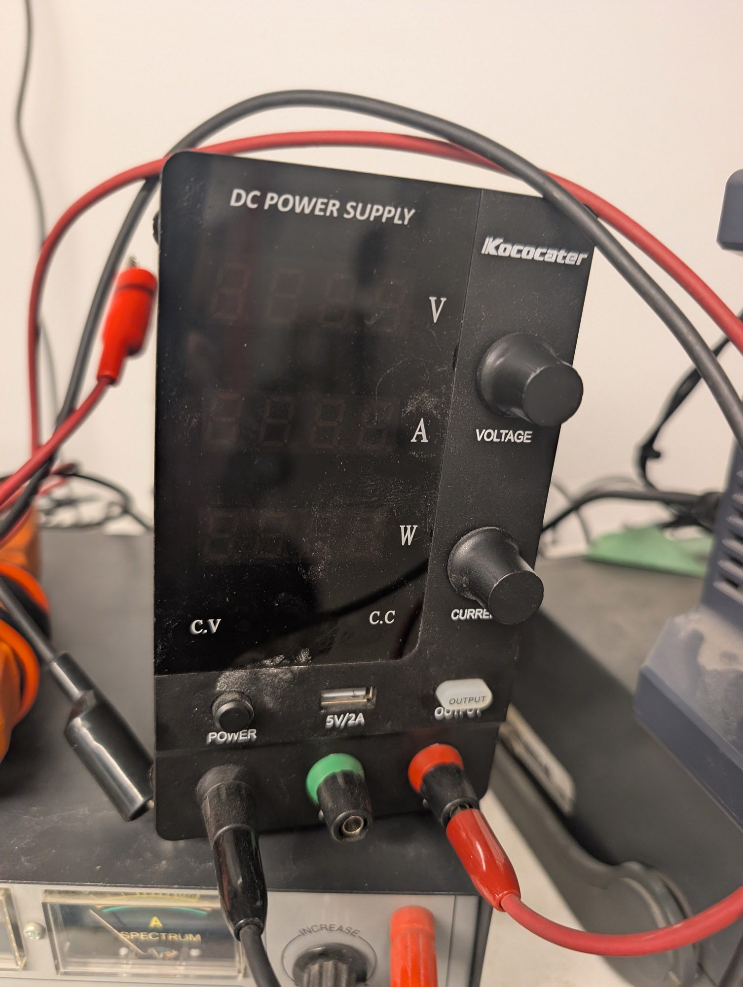 A black rectangular power supply, about twice as tall as it is wide. It says “IKocater” on the top right and “DC Power Supply” at the top. The right side has Voltage and Current dials, the left side has four digit 7-segment displays for V, A, and W, and LEDs for CV and CC. Black, green, and red combo banana/binding posts are at the bottom.